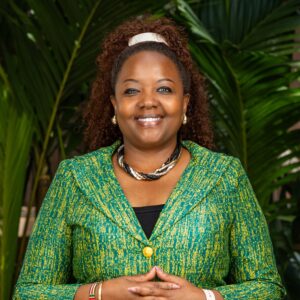Winnie Wambugu, a black lady with red-brown long, curly hair in a green jacket, smiling at the camera.
