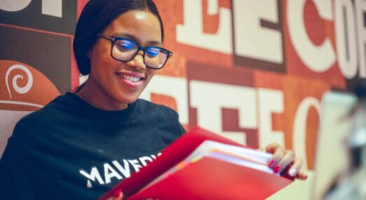 Madonna Seroto, owner of Maverick Coffee, stands in her coffee shop and reviews her daily itinerary file in Pretoria, South Africa. She is a young Black woman with glasses and her straight hair tied black, wearing a black t'shirt and smiling.
