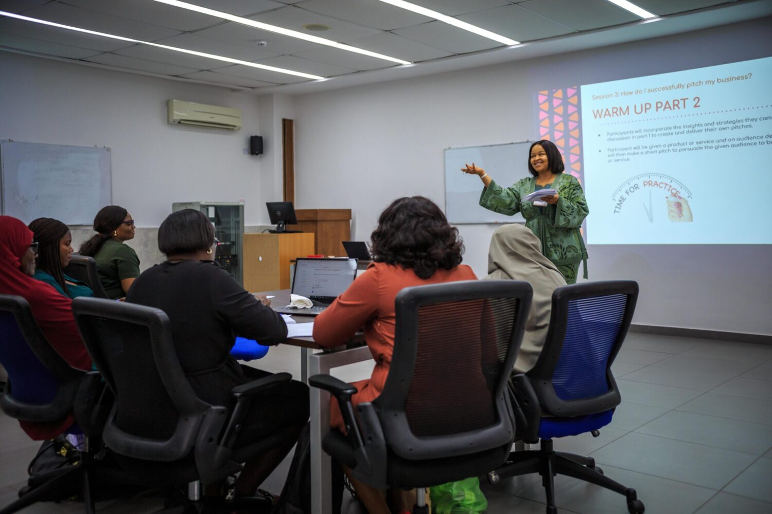 A group of women sit in a classroom listening to a Road to Finance trainer who is presenting at the front of the class.