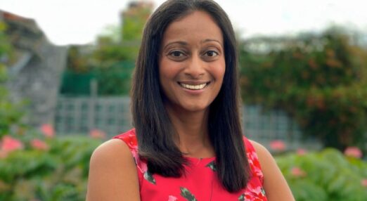 Dhivya O'Connor, a woman of South Asian heritage with medium-length straight, dark brown hair, is smiling at the camera. She is standing in a garden wearing a red dress.