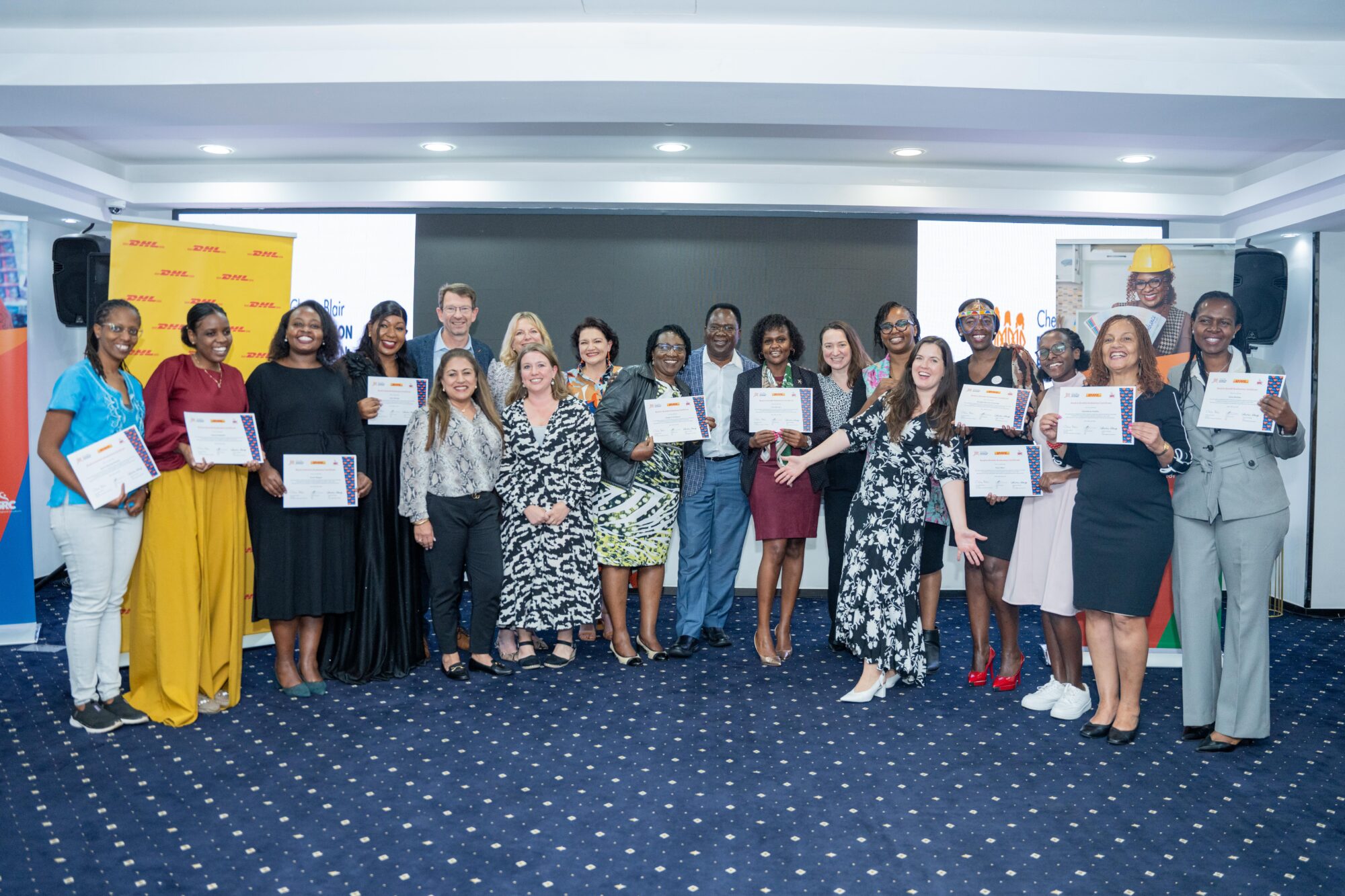 A group of around 20 people standing posed together in a conference-style space. Some are holding certificates and all are smiling at the camera.