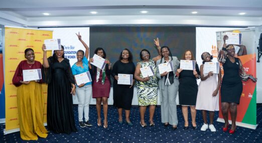 A group of Kenyan women stand in a line in a conference-style room. They are smiling and holding certificates.