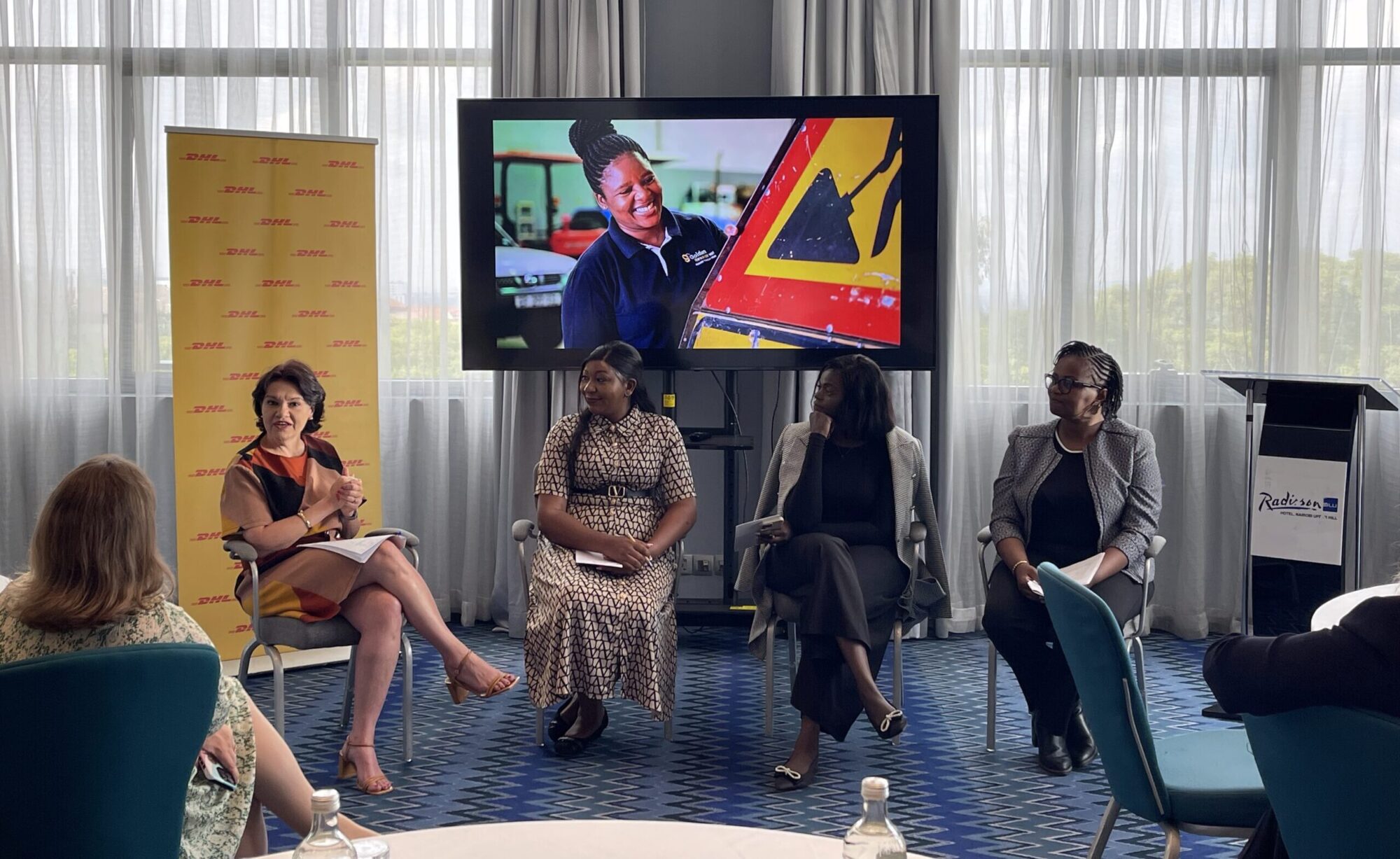 Four women sit together on a panel in a conference-style room. There are audience members facing the women.