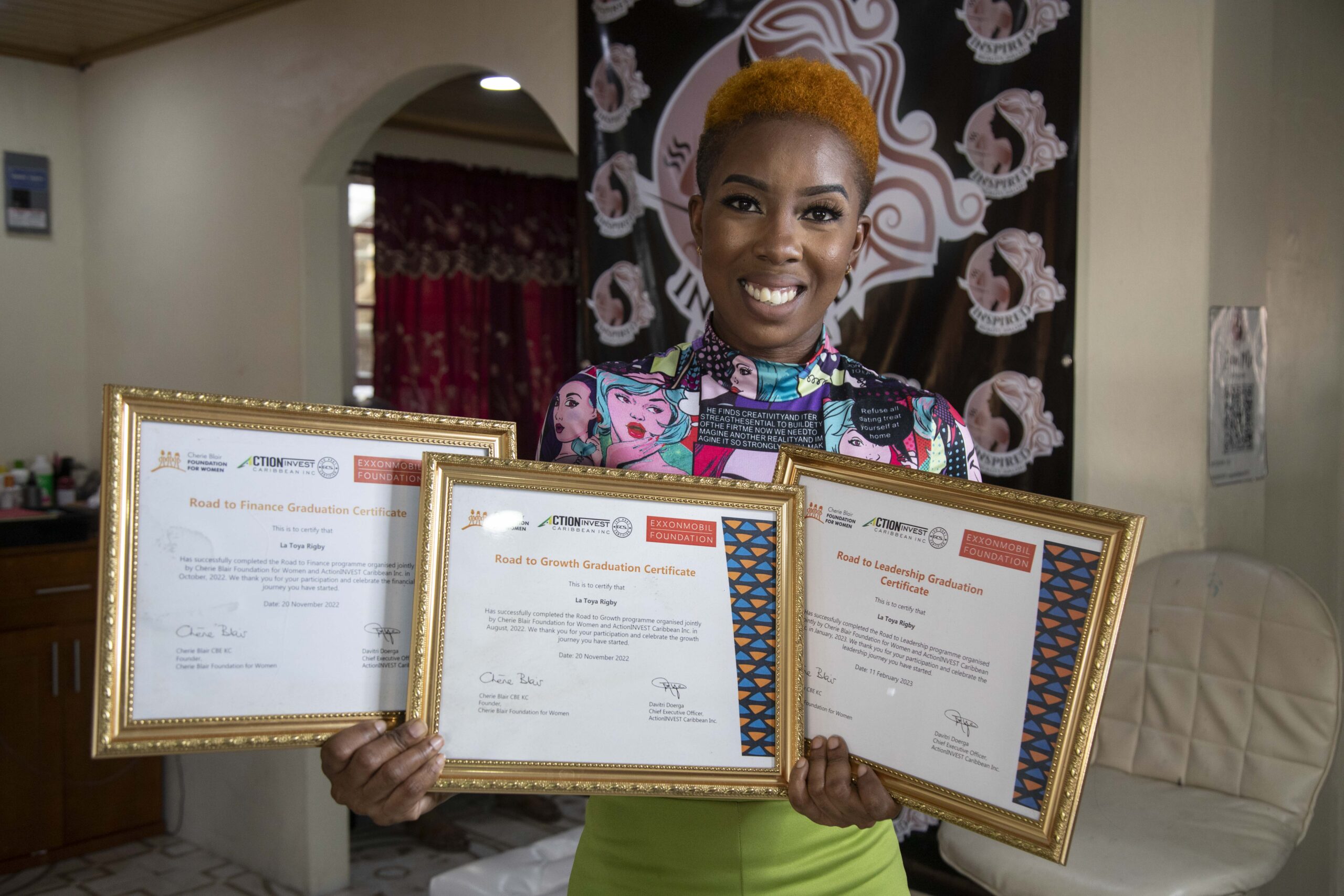 Latoya Rigby, a Black woman with very short, orange hair, poses in a brightly coloured patterned top and bright green trousers. She is smiling and holding three framed training certificates.