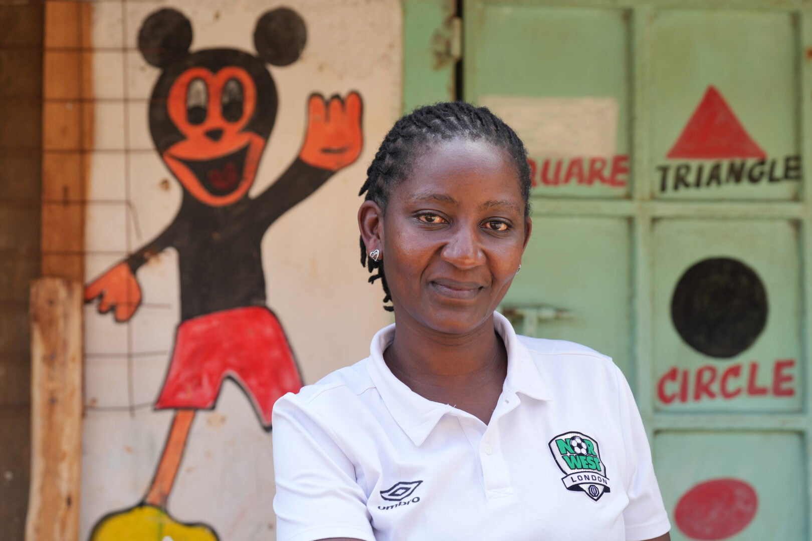 Mary Giroga, a woman entrepreneur in Kenya smiles in front of a wall with shape blocks painted onto it. She is wearing a white polo shirt.