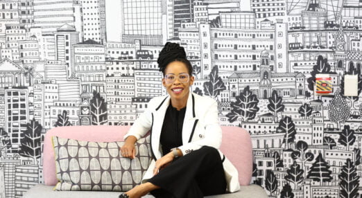Queen Mokulubete, a Black South African woman with black hair in long braids up in a bun and glasses, She is wearing a white blazer, black blouse and black trousers. She is smiling as she poses on a pink sofa in her office in front of a wall covered in black and white line drawings.