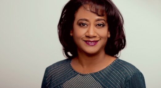 Head and shoulders portrait of Tanya Joseph, in front of a plain white background. She is a Black woman with shoulder-length dark brown hair and is wearing purple lipstick and a smart grey top. She is smiling at the camera.