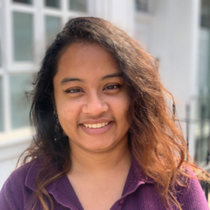 Aradhana Choudhury smiles in a purple top. She has wavy brown hair which gleams red in the sunlight. She is outside in front of a door and some windows.