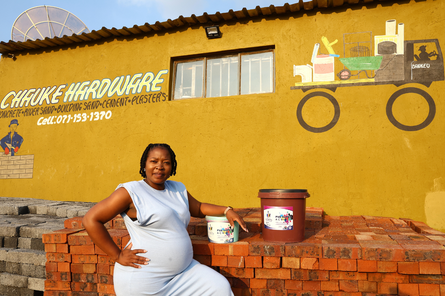 Zandile Manyisa, a woman entrepreneur in South Africa, poses in front of a bright yellow hardware store. She is leaning on a tub of paint.