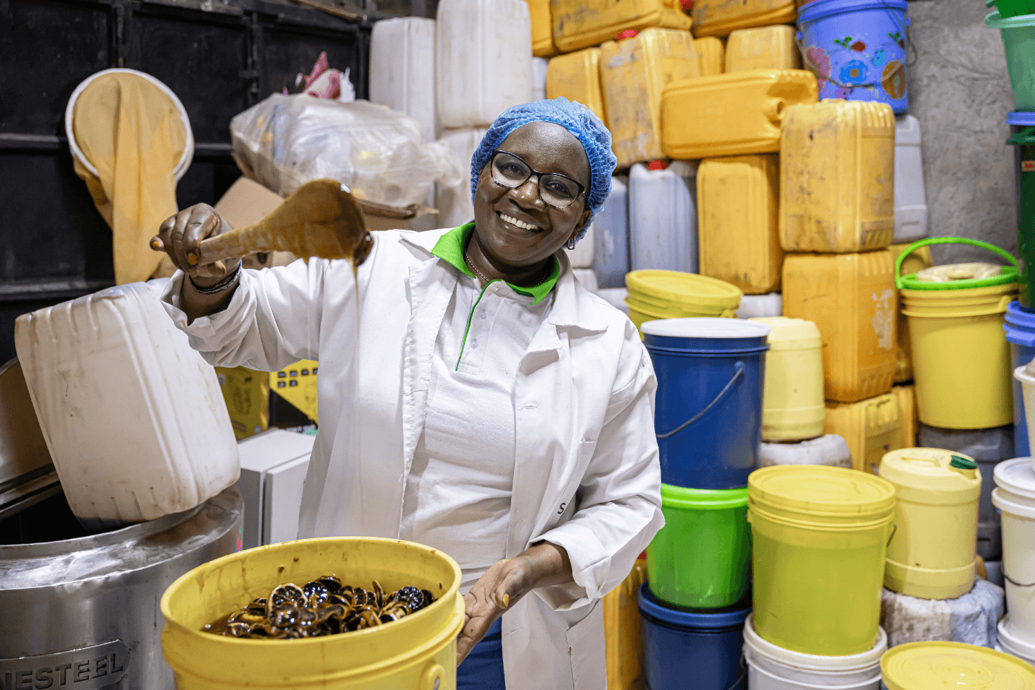 Margaret Mwuara, a woman entrepreneur from Kenya, is standing with a large plastic container full of honey. She is holding a wooden spoon, dripping with honey, above the container.