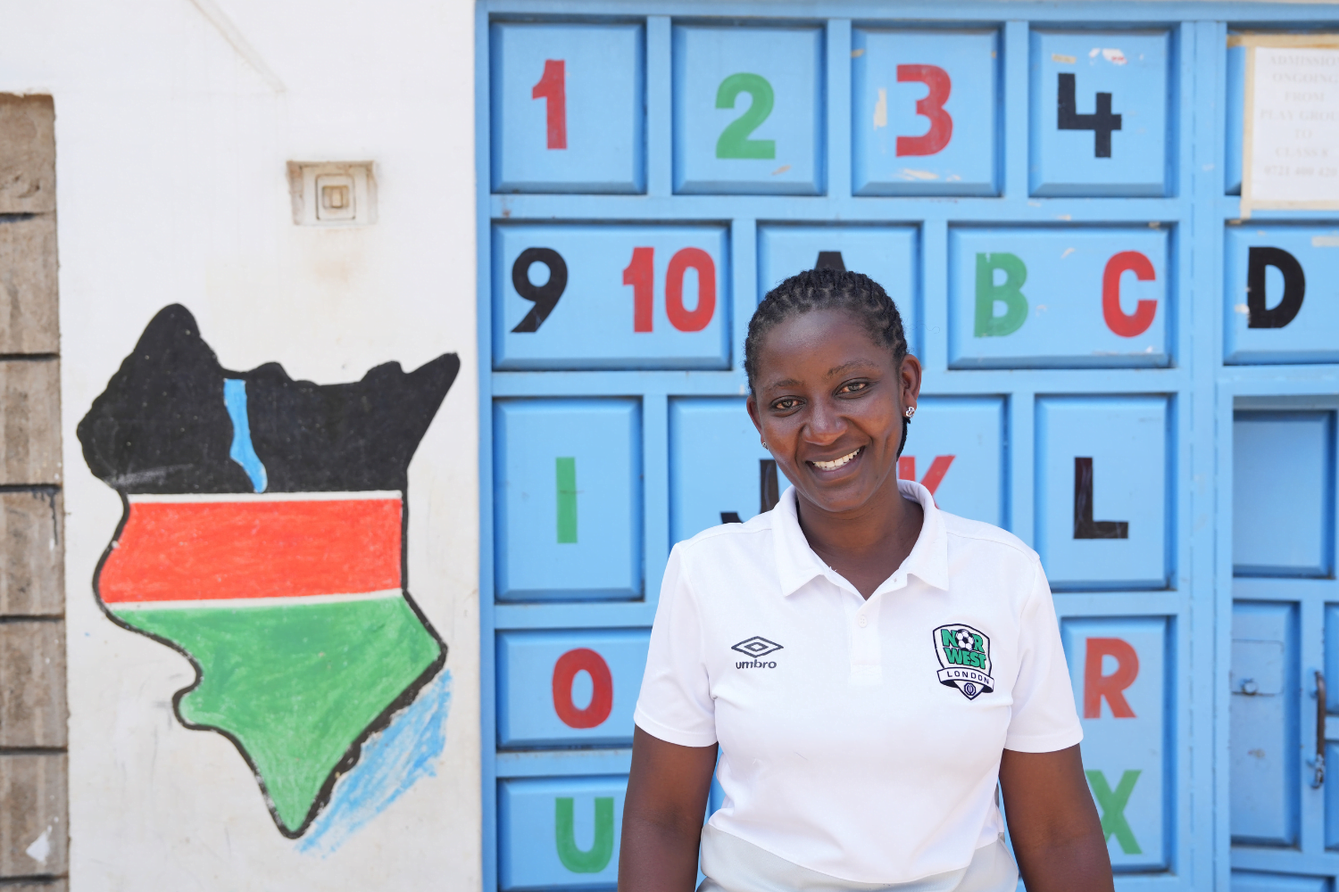 Mary Giroga, a woman entrepreneur in Kenya smiles in front of a wall with number and letter blocks painted onto it. She is wearing a white polo shirt.