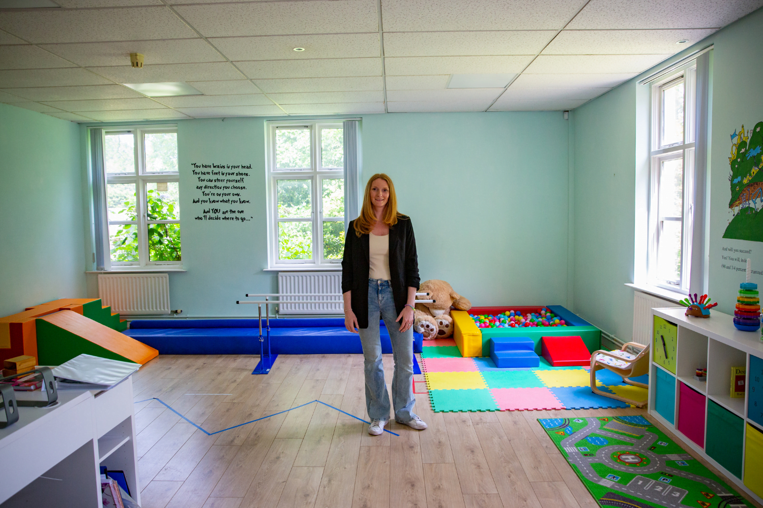 Sam Tebb, a woman entrepreneur from the UK, stands in the middle of a playroom. There is a ball pit in the corner and lots of bright primary colours. She is wearing jeans and a white shirt with a black blazer over top.