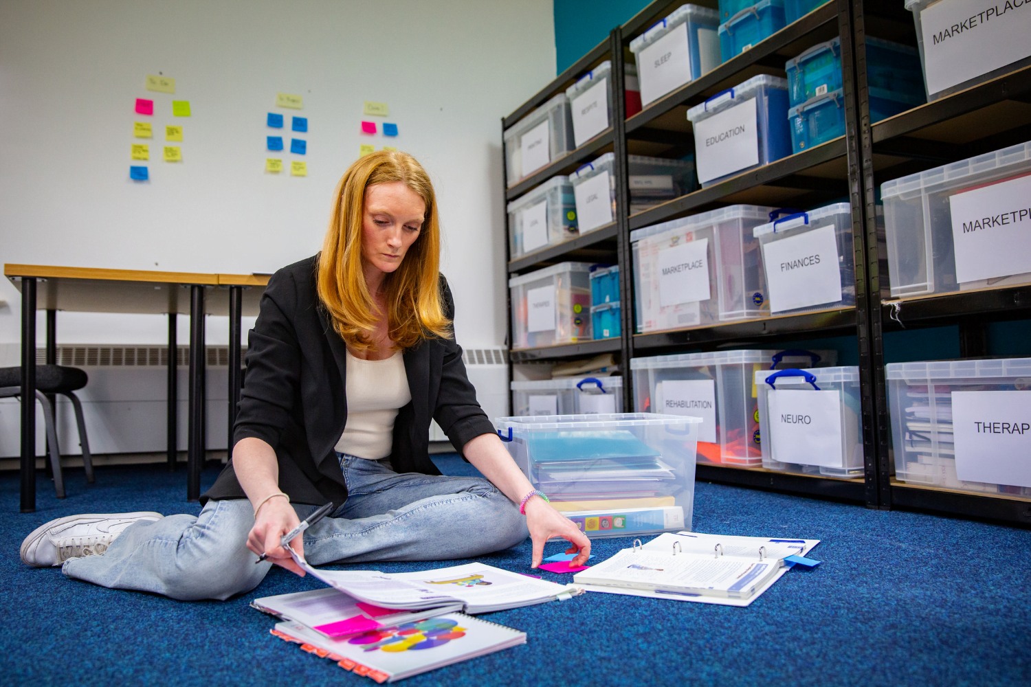 Woman entrepreneur Sam Tebb sits on the floor. To her left, a large storage unit labelled with things like 'Education', 'Finances' and 'Neuro' is against the wall. She is surrounded by three books, which she is labelling with brightly coloured sticky notes.