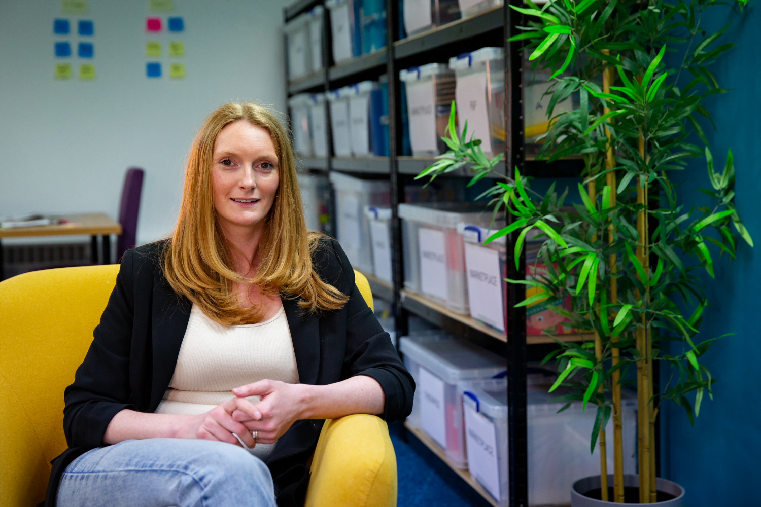 Sam Tebb, a woman entrepreneur from the UK, sits in a yellow armchair. She is wearing jeans and a white shirt with a black blazer over top. Next to her is a small potted tree and behind her are containers with informative documents for parents of disabled children.
