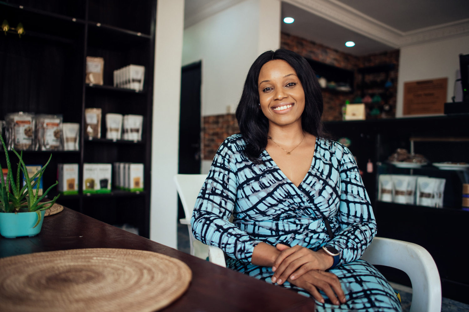 A woman entrepreneur from Nigeria sits at a wooden table in a white chair. She leans her crossed arms on the chair. She is smiling and wearing a blue patterned dress. Her hair is down.