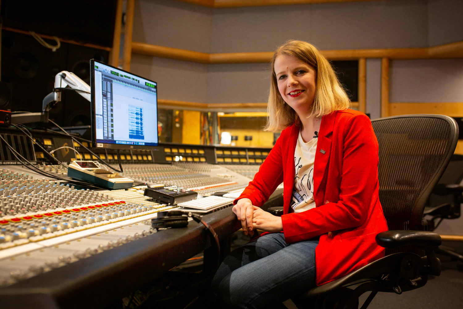 Olga FitzRoy, a British self-employed sound engineer, sits at a sound desk with hundreds of buttons on it. She is smiling at the camera.