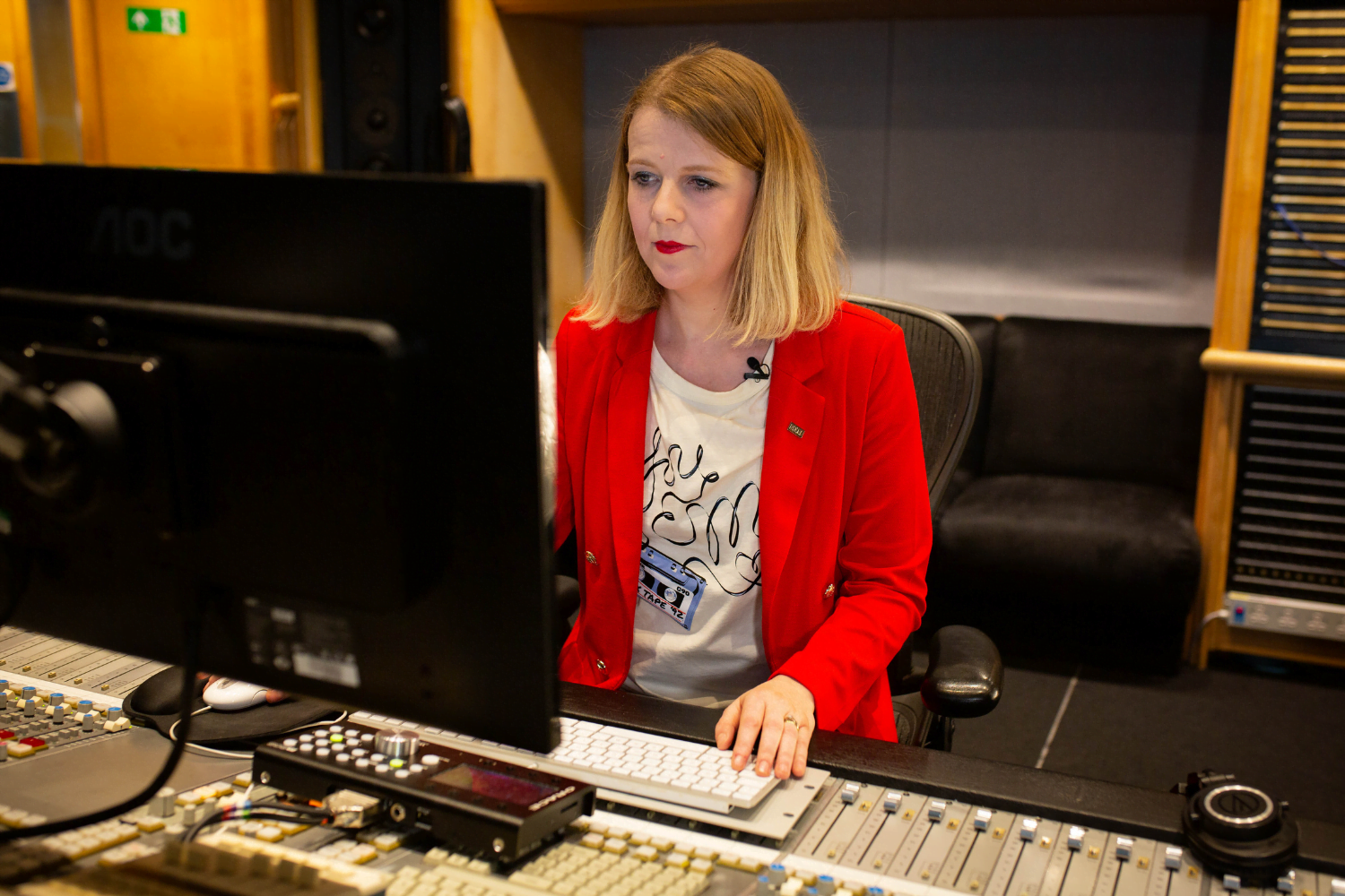 Olga FitzRoy, a British self-employed sound engineer, sits at a sound desk with hundreds of buttons on it. She is looking at a desktop computer.