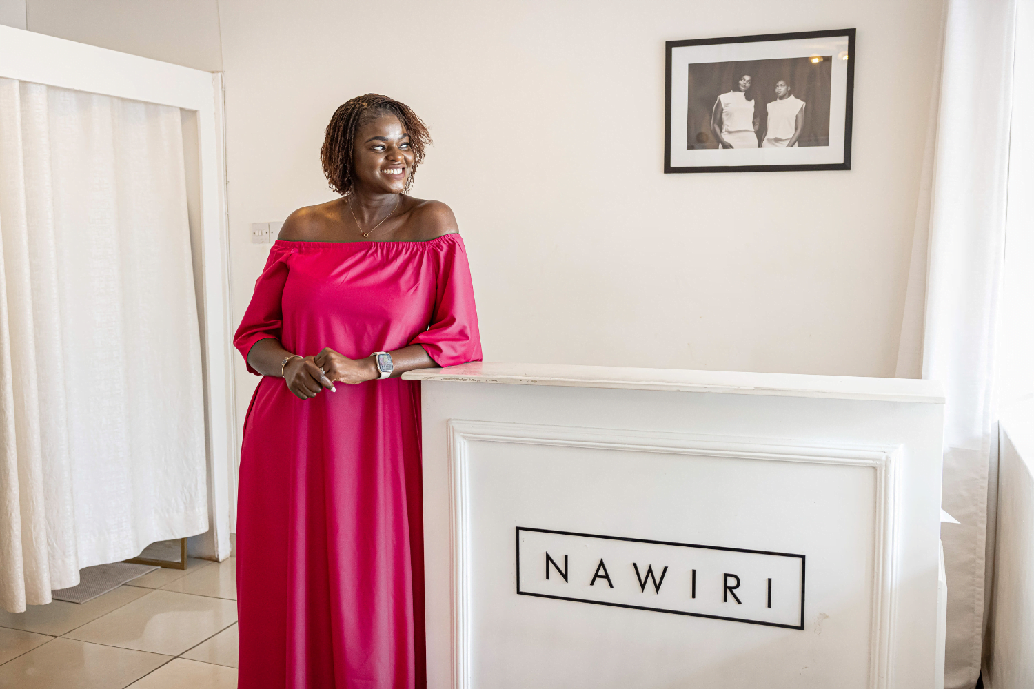 Joy Obuya, a woman entrepreneur and fashion designer from Kenya, smiles into the distance while leaning sideways against the counter of her clothing store. She is wearing a magenta, off-shoulder dress that is floor length. Her hair is jaw-length and curly. In the background is a white curtained dressing room.