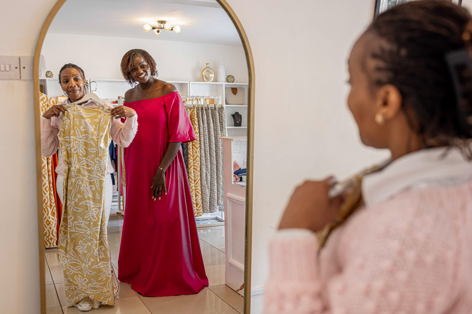 Joy Obuya, a woman entrepreneur and fashion designer from Kenya, works with a customer in her clothing store. The customer is holding up a patterned floor length dress in front of a long arched mirror. Joy stands beside her in a floor length magenta dress. In the foreground you can see the shoulder and side profile of the customer as she looks at their image in the mirror.