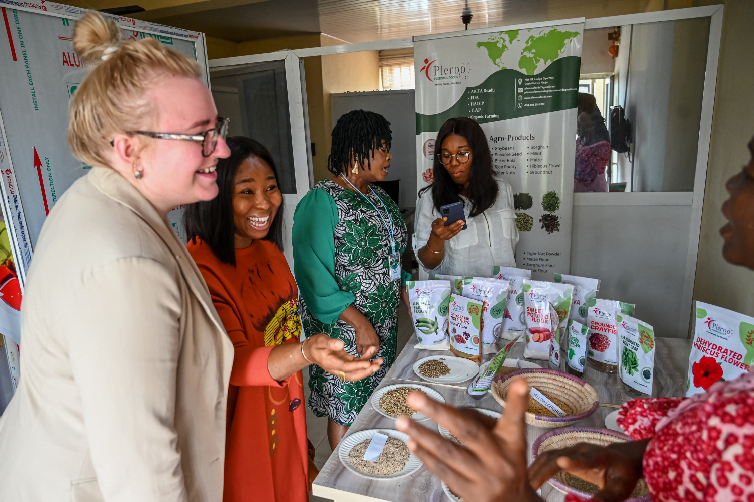 Nigerian mentee Ellah Omezi and American mentor Gwynneth Anderson stand around a table of Nigerian produce, both talking to other women also stood around the table.