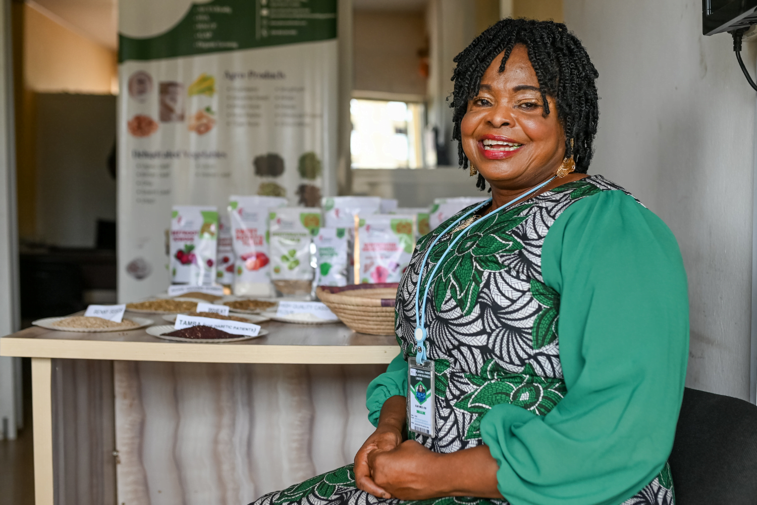 Ellah Omezi, a Nigerian woman entrepreneur wearing a green embroidered dress, grins at the camera in front of some labelled seeds on a table behind her.