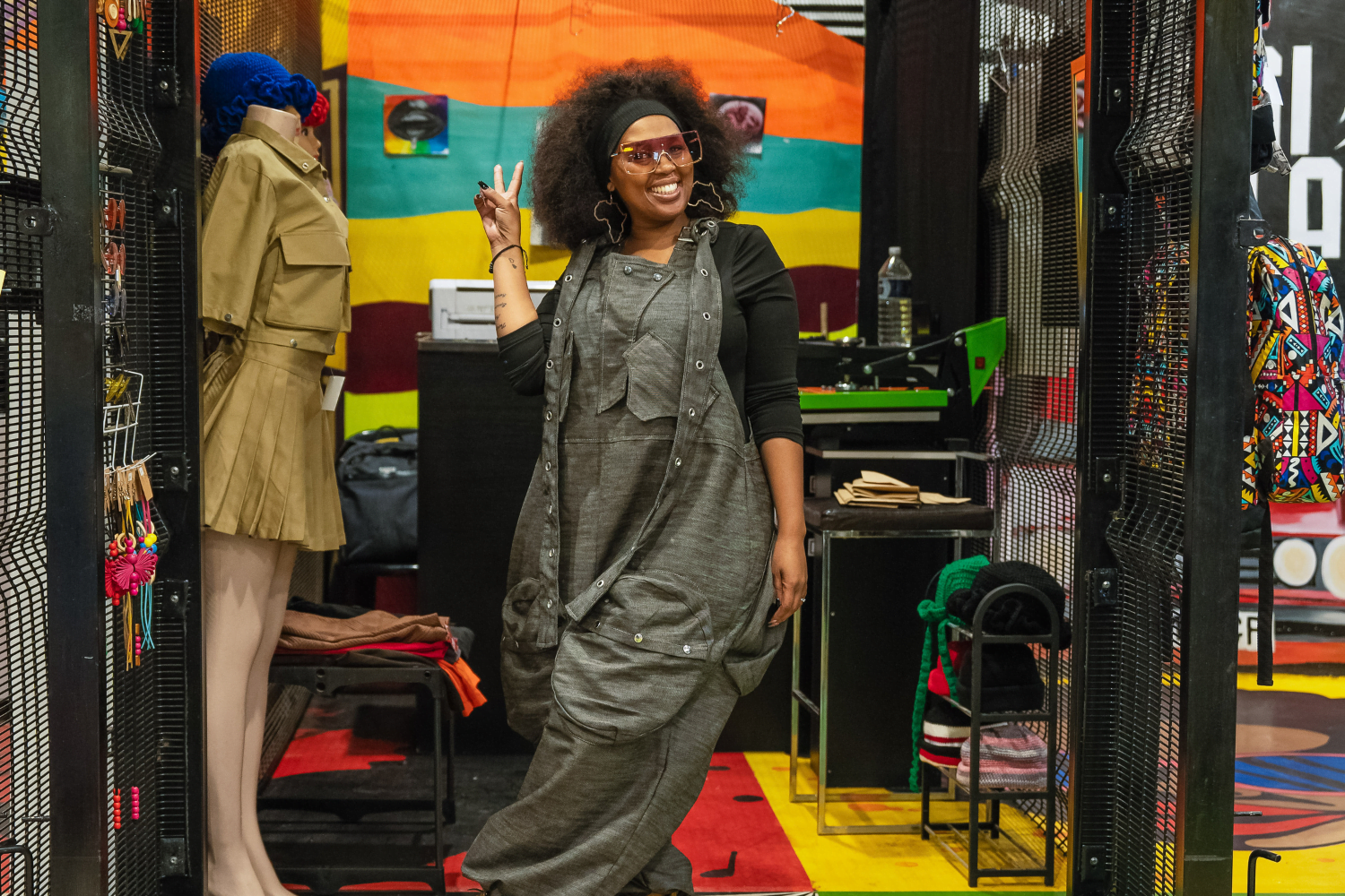 Nkeletseng Tsetsane, a woman entrepreneur in South Africa poses in her store front. She has one leg crossed in front of the other and is giving a peace sign. She is wearing grey dungarees with a black long sleeved top underneath. Her hair has a black headband in it and she has lightly tinted sunglasses on. Next to her is a mannequin with a tan coordinated skirt and top set. Behind her there seems to be garment manufacturing equipment.
