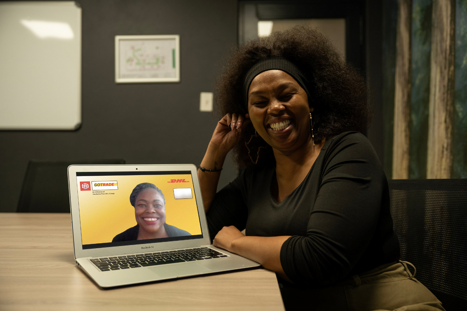 Nkeletseng Tsetsane, a woman entrepreneur in South Africa, smiles as she leans her arms against a wooden table. In front of her on the wooden table is a laptop with a woman on the screen. This is her mentor, Glory Mutai from Kenya. The two of them are facing the camera and smiling.