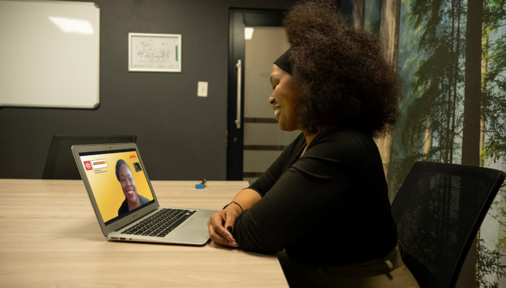 Nkeletseng Tsetsane, a woman entrepreneur in South Africa, smiles as she leans her arms against a wooden table. In front of her on the wooden table is a laptop with a woman on the screen. This is her mentor, Glory Mutai from Kenya. The two of them are smiling at each other.