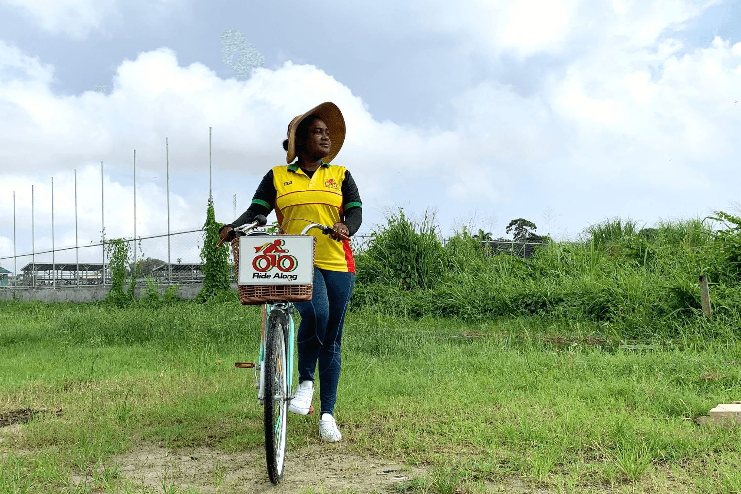 Cherry-Ann Greene, a woman entrepreneur from Guyana, is smiling. She is also holding a bicycle by the handlebars. Her bike has a basket with 'a Ride Along logo' printed on it. The same logo is printed on her shirt.