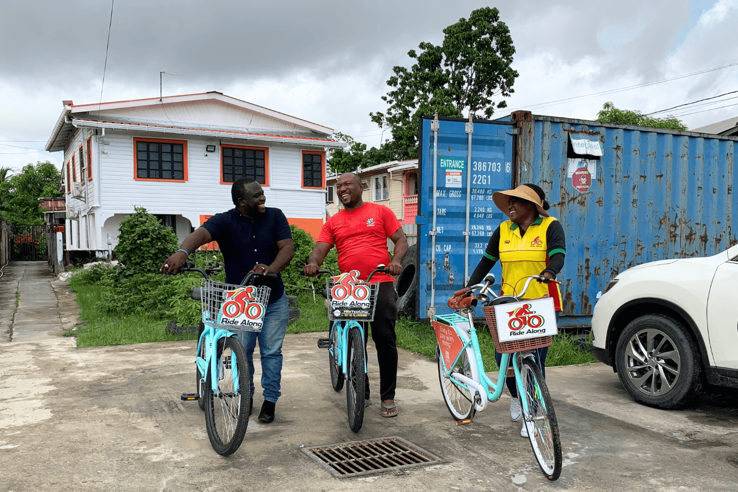 Cherry-Ann Greene, a woman entrepreneur from Guyana, is holding a bicycle by the handlebars. Her bike has a basket with 'a Ride Along logo' printed on it. The same logo is printed on her shirt. She is smiling at two men next to her who also hold bicycles with baskets with the 'Ride Along' logo printed on them.