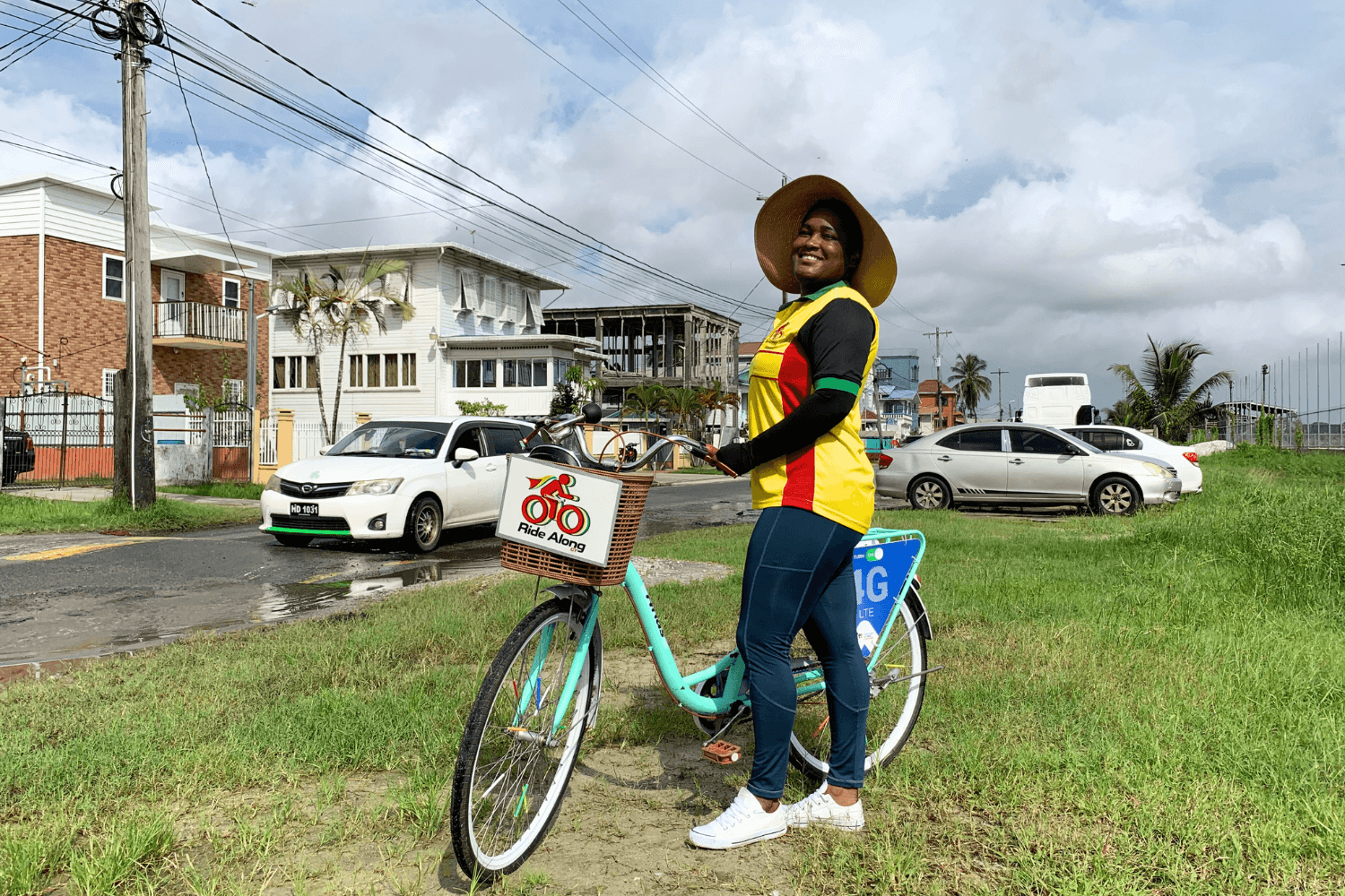 Cherry-Ann Greene, a woman entrepreneur from Guyana, is smiling at the camera and holding a bicycle by the handlebars. Her bike has a basket with 'a Ride Along logo' printed on it. The same logo is printed on her shirt.