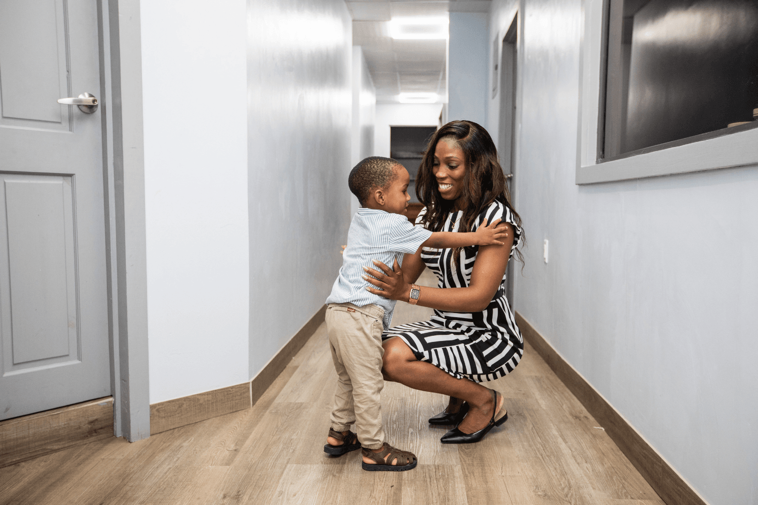 Candace Wickham crouches down to embrace her toddler son. Candace is wearing black heels and a white and black patterned dress. Her hair is down and wavy. Her son is wearing chinos, black shoes, and a light blue button down shirt. They are both Afro-Guyanese. They are standing inside in a corridor.