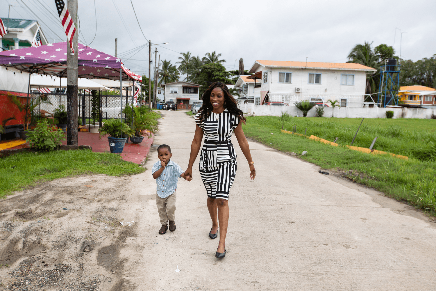 Candace Wickham and her young son walk down a quiet street holding hands. Candace is wearing black heels and a white and black patterned dress. Her hair is down and wavy. Her son is wearing chinos, black shoes, and a light blue button down shirt. They are both Afro-Guyanese. On the right had side of the photo in the background there is a white house with an orange roof. On the left side, there is a purple sun umbrella shading a red brick driveway. In the far background, there are some palm trees popping out from behind a house.