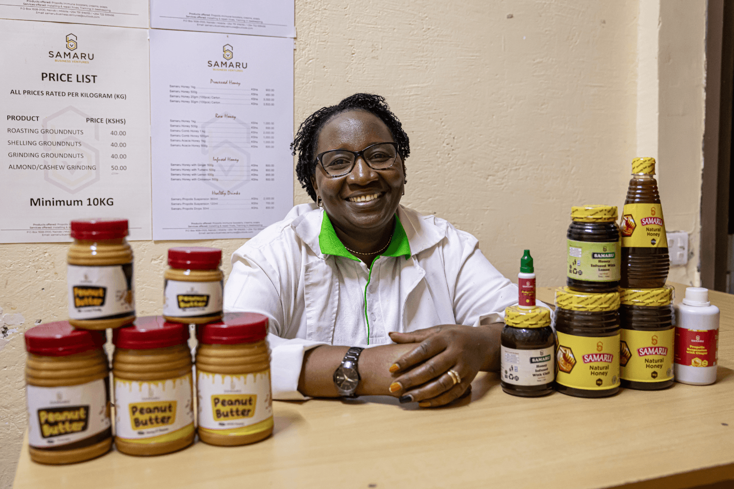 Margaret Mwuara, a woman entrepreneur from Kenya, sits behind a table with her produce (honey and peanut butter) piled on it. She is smiling at the camera.