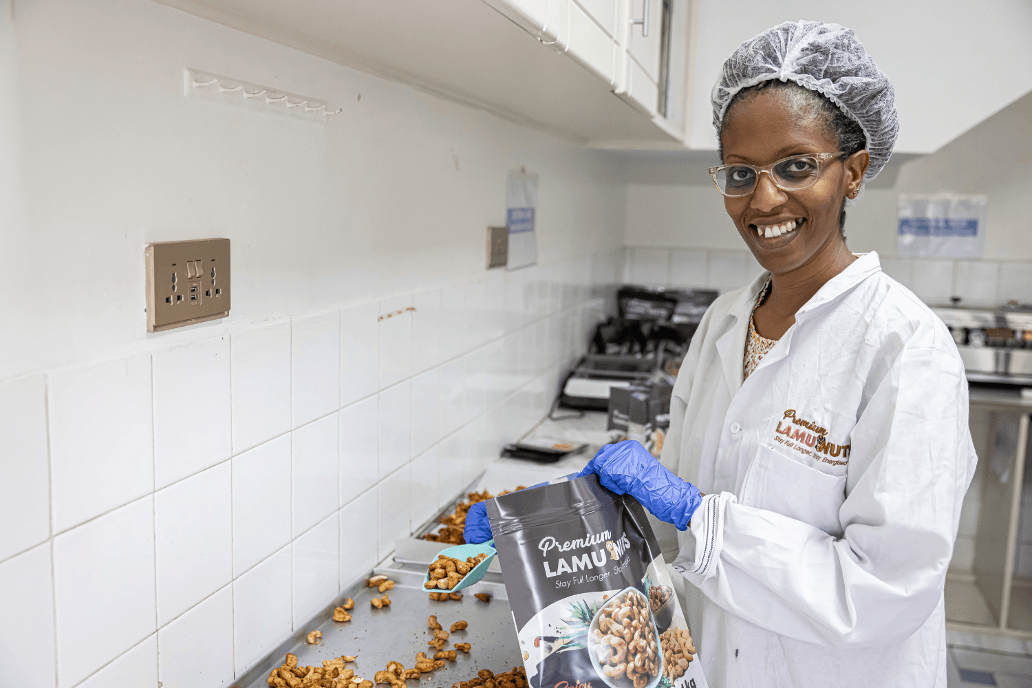 Naomi Kimuyu, a woman entrepreneur from Kenya, is standing in a kitchen smiling at the camera. She is wearing a hairnet, latex gloves, and a white chef's jacket with a 'Premium Lamu Nuts' logo. She is standing in front of a tray of roasted, seasoned nuts, and is using a scoop in her right hand to scoop the nuts from the tray into a bag in her left hand. The bag is also printed with a 'Premium Lamu Nuts' logo.