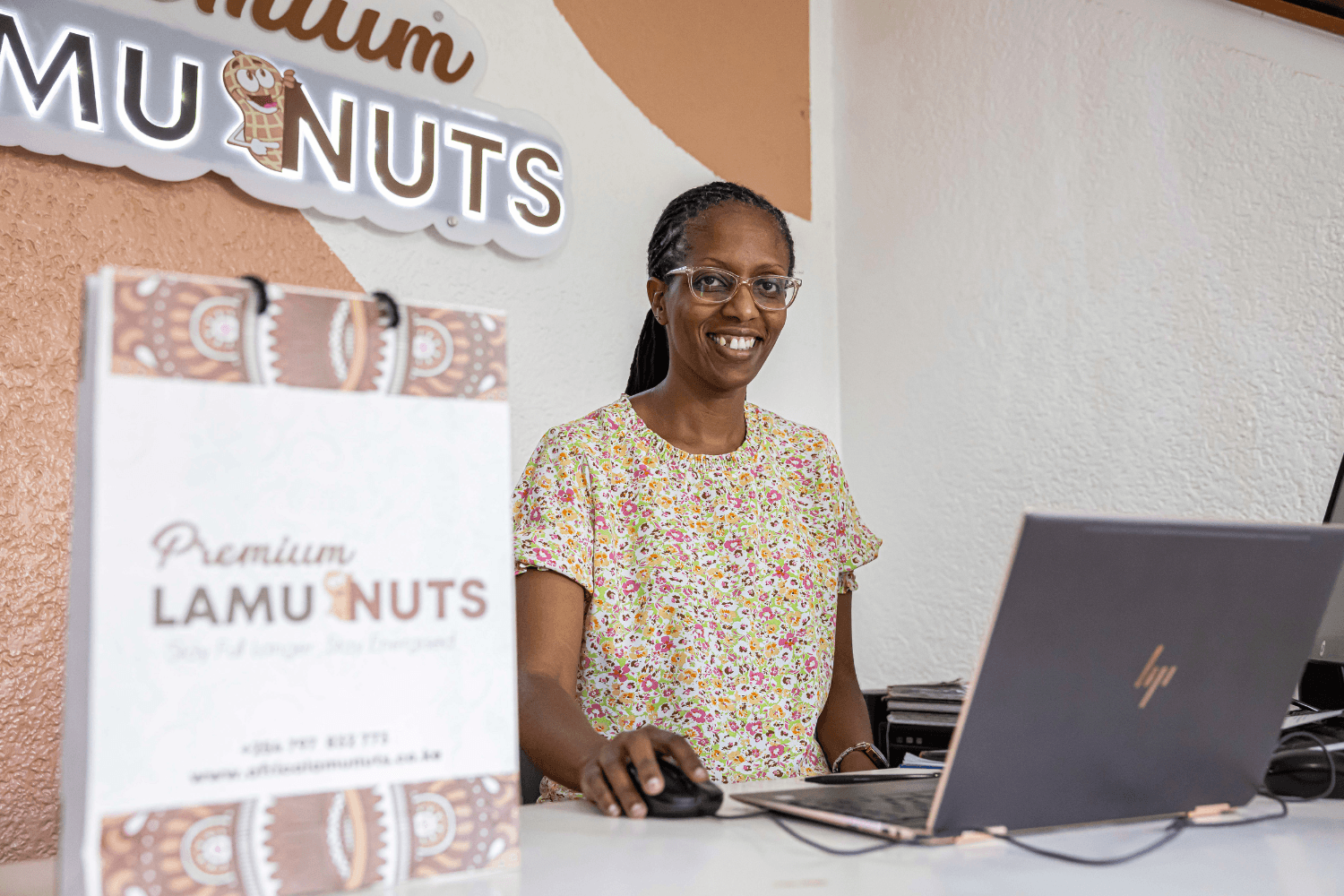 Naomi Kimuyu, a woman entrepreneur from Kenya wearing a floral top and glasses, is sat at a desk working on a laptop. Beside her on the desk is a gift bag with a'Premium Lamu Nuts' logo. The same logo is on the wall behind her.