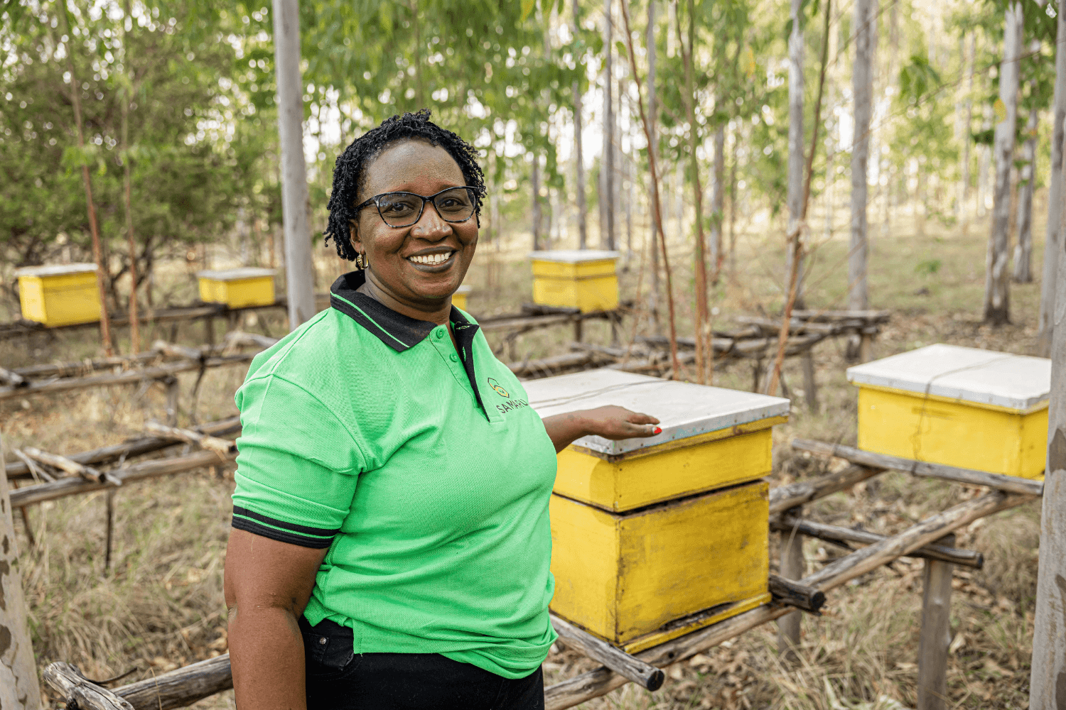 Margaret Mwuara, a woman entrepreneur from Kenya, stands smiling at the camera with her hand on a beehive. Rows of other beehives are visible behind her. She is wearing a green polo shirt with 'Samaru' logo on it.