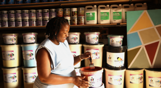 Zandile Manyisa, a woman entrepreneur in South Africa, smiles. She is holding a paint brush in her hand, which shines with red paint from a tub of paint beside her.