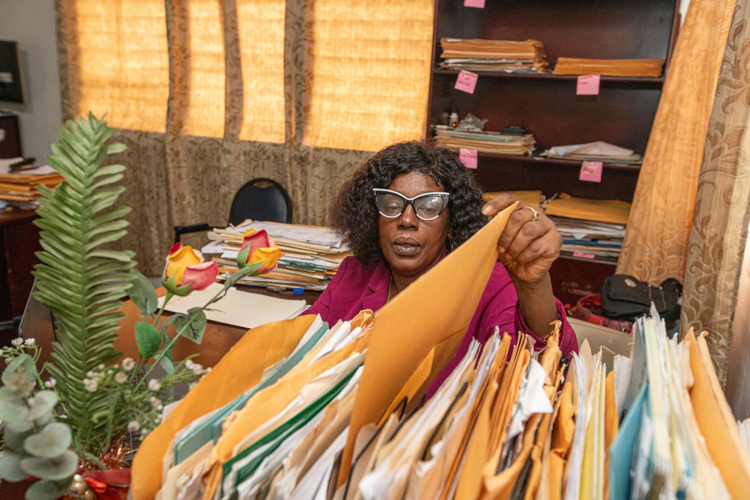 Dionne Thomas, a woman entrepreneur from Guyana, sits behind a dark wooden desk inside her office. She is reaching up toward the camera to put a file on a shelf full of files, which are in the foreground of the image. Dionne is wearing glasses with black and white frames, a magenta blazer over a black shirt, and a gold necklace.