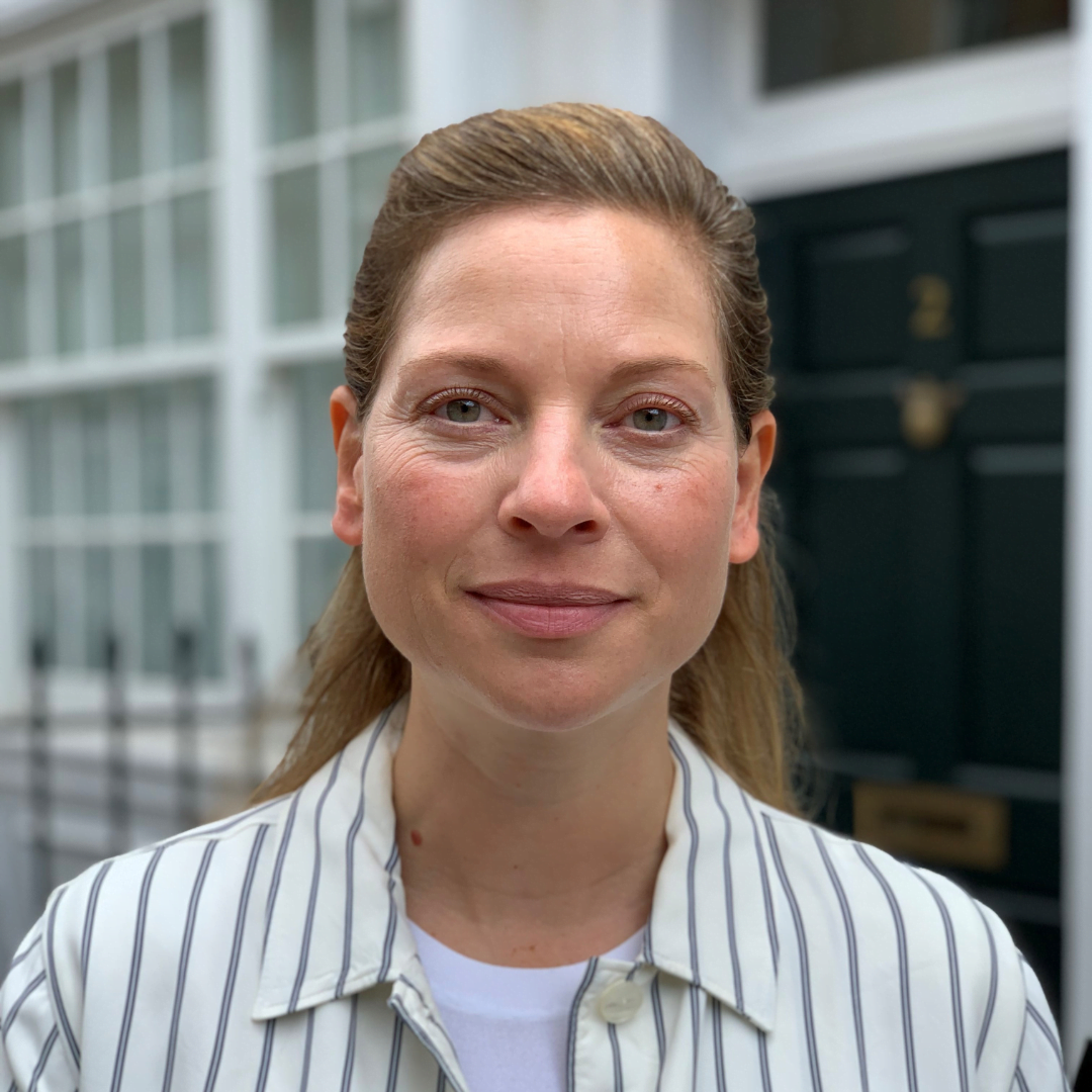 Bree Dellepoort-Walsh smiles in a striped button down shirt. Her blonde hair is pinned in a half-up, half-down hairstyle. She is outside in front of a door and some windows.