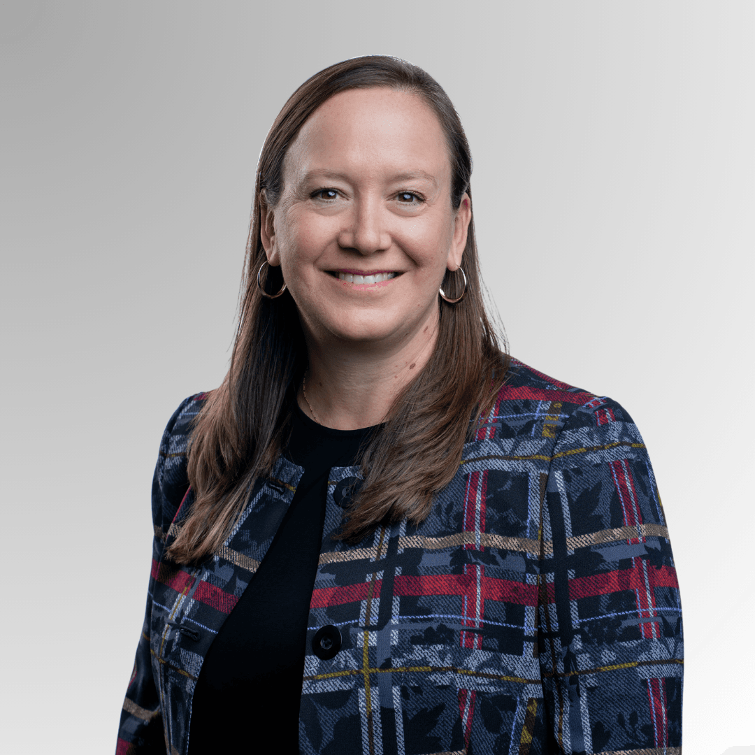 Suzanne Ehlers smiles in a blue and pink plaid blazer over a black shirt. She is a white woman with medium length brown hair.