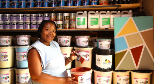 Zandile Manyisa, a woman entrepreneur in South Africa, smiles at the camera. She is holding a paint brush in her hand, which shines with red paint from a tub of paint beside her.