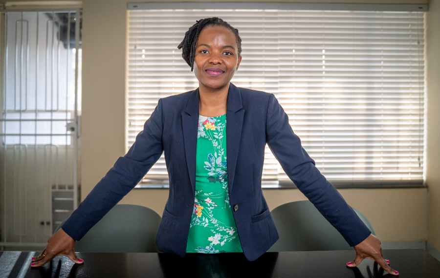 Dudu Makhari, a woman entrepreneur from South Africa, poses confidently in her office. She is standing behind a wooden desk with her hands spread far apart as she leans against it. She is wearing a blue blazer and a green floral shirt. Her hair is tied up and she is smiling.