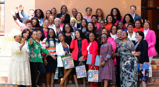 A group of women in South Africa pose for a group photo on some grass in front of an orange building. Many are holding bags and certificates from their graduation ceremony.