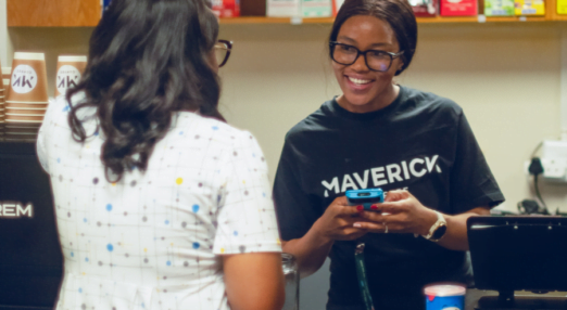 Madonna Seroto, Owner of Maverick Coffee in South Africa, smiles at a customer during a transaction in her coffee shop. The customer has her back turned to the camera and is wearing a white dress with blue and grey dots on it. Madonna has her hair tied back in a ponytail. She is wearing a shirt that says "Maverick" on it and holding a smartphone, which she's using for the transaction.