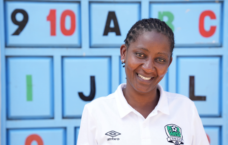 Mary Giroga, a woman entrepreneur in Kenya smiles in front of a wall with number and letter blocks painted onto it. She is wearing a white polo shirt.