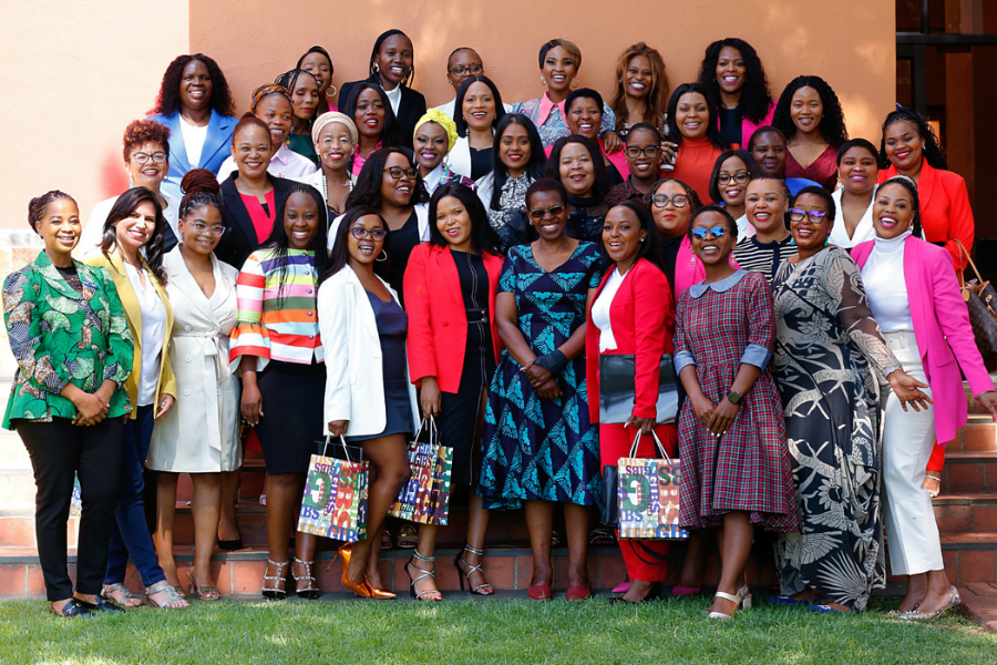 A group of women in South Africa pose for a group photo on some grass in front of an orange building. Many are holding bags and certificates from their graduation ceremony.