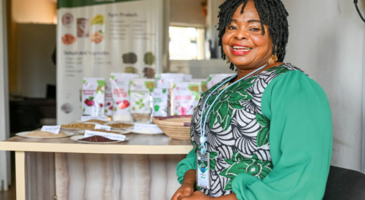 Ellah Omezi, a Nigerian woman entrepreneur wearing a green embroidered dress, grins at the camera in front of some labelled seeds on a table behind her.
