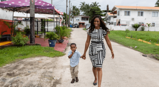 Candace Wickham and her young son walk down a quiet street holding hands. Candace is wearing black heels and a white and black patterned dress. Her hair is down and wavy. Her son is wearing chinos, black shoes, and a light blue button down shirt. They are both Afro-Guyanese. On the right had side of the photo in the background there is a white house with an orange roof. On the left side, there is a purple sun umbrella shading a red brick driveway. In the far background, there are some palm trees popping out from behind a house.